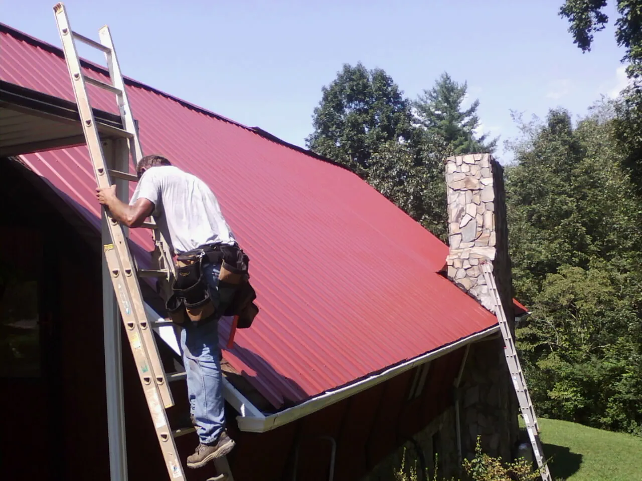 Roofer on a ladder beside a red metal roof and stone chimney — Soffit & Fascia Repair by Roofing Brothers in Channelview
